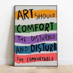 Op de afbeelding: Een zwart ingelijste poster met een witte achtergrond en kleurrijke strepen. De tekst op de poster luidt "Kunst moet de verstoorden troosten en de comfortabelen verstoren" met de toeschrijving "@dormousestudio".