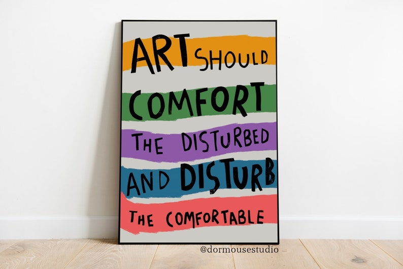 Op de afbeelding: Een zwart ingelijste poster met een witte achtergrond en kleurrijke strepen. De tekst op de poster luidt "Kunst moet de verstoorden troosten en de comfortabelen verstoren" met de toeschrijving "@dormousestudio".