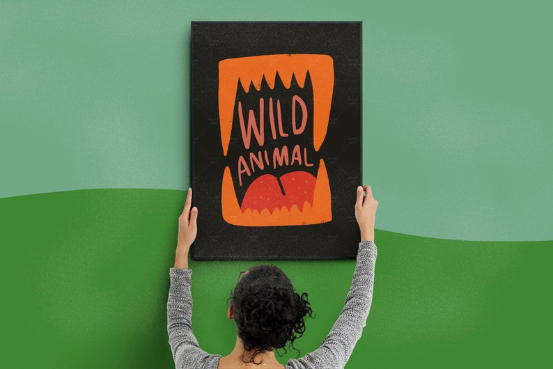 May include: A person hangs a black canvas print with an orange and red illustration of a mouth with sharp teeth and the text "WILD ANIMAL" on a green wall.