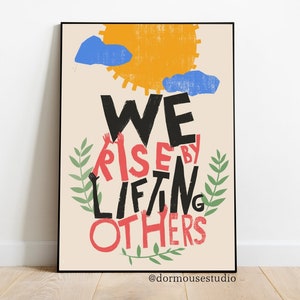 Op de afbeelding: Een beige poster met een zwarte lijst toont een kleurrijke illustratie van een zon, wolken en groene bladeren. De tekst "WE RISE BY LIFTING OTHERS" is geschreven in zwarte en rode letters, waarbij de woorden "RISE" en "OTHERS" rood zijn.