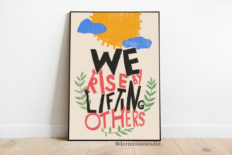 Op de afbeelding: Een beige poster met een zwarte lijst toont een kleurrijke illustratie van een zon, wolken en groene bladeren. De tekst "WE RISE BY LIFTING OTHERS" is geschreven in zwarte en rode letters, waarbij de woorden "RISE" en "OTHERS" rood zijn.