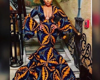 May include: A long, patterned dress with a deep neckline and flared sleeves. The dress features a vibrant geometric design in blue, yellow, and orange. The dress has a mermaid silhouette and is worn on a staircase.