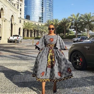 May include: A woman wearing a gray and brown patterned dress with a fitted bodice and a full skirt. The dress has a wide, bell-shaped sleeve and a belt at the waist. She is wearing black high heels and sunglasses.