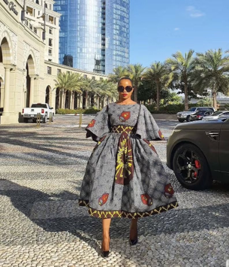 May include: A woman wearing a gray and brown patterned dress with a fitted bodice and a full skirt. The dress has a wide, bell-shaped sleeve and a belt at the waist. She is wearing black high heels and sunglasses.