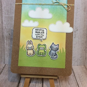May include: A handmade card with a yellow, green, and blue watercolor background. The card features three animals wearing face masks: a rabbit, a frog, and a cat. The card also includes a speech bubble with the text "sending SMILES with my EYES".