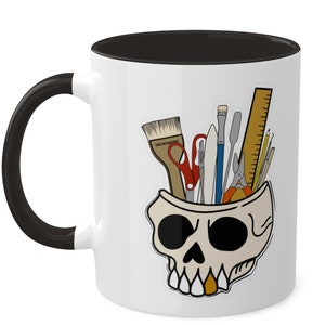 May include: White ceramic mug with a black handle and black rim. The mug features a graphic of a skull with various art supplies inside, including paintbrushes, scissors, a ruler, and a pencil.