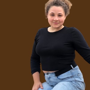 May include: A woman with curly brown hair is wearing a black long-sleeved shirt and blue jeans. She is smiling and looking at the camera.