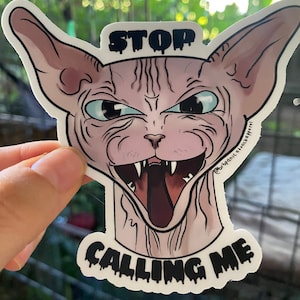 May include: A sticker featuring a cartoon Sphynx cat with a menacing expression. The cat has a large open mouth with sharp teeth and a long tongue. The sticker has the text "STOP CALLING ME" in dripping black letters.