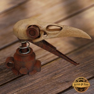 Könnte beinhalten: Eine Steampunk-Skulptur eines Vogelschädels mit langem Schnabel, montiert auf einem rostigen Metallsockel mit Feder. Der Schädel ist cremefarben, die Metallteile in Braun- und Rottönen gehalten. Die Skulptur steht auf einer Holzoberfläche.