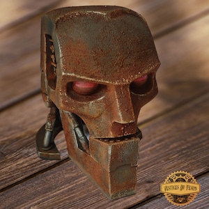 Puede incluir: Una escultura de cabeza de robot metálica y desgastada con ojos rojos brillantes. La escultura tiene un aspecto oxidado y envejecido, con detalles mecánicos visibles y una mandíbula cuadrada. La pieza está sobre un fondo de madera, evocando una estética vintage o steampunk.