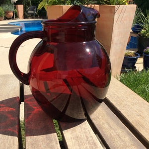 Puede incluir: Una jarra de vidrio roja grande y redonda con asa. La jarra está sobre una mesa de madera.