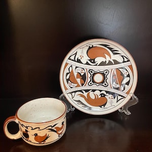 May include: A ceramic cup and saucer set with a hand-painted design. The cup and saucer feature a cream-colored base with brown and black bird and geometric patterns. The cup has a brown handle and rim.