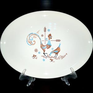 May include: An oval, white ceramic platter with a mid-century modern design. The platter features a brown and blue rooster and hen illustration with floral and geometric accents. The platter is displayed on a clear stand.