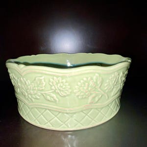 May include: A light green ceramic bowl with a floral and diamond pattern. The bowl has a scalloped rim and is likely used for decorative purposes or serving food. The design is embossed, adding texture to the surface.