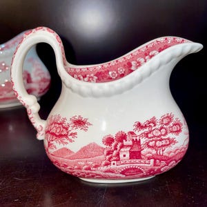 Puede incluir: Cremera de cerámica blanca con un diseño de paisaje rojo que presenta un castillo, un puente y árboles. La cremera tiene un asa curva y un patrón floral decorativo alrededor del borde. Una pieza de vajilla clásica.