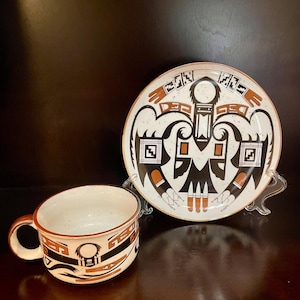 May include: A cream-colored cup and plate set with a Native American-inspired design. The plate and cup feature black, brown, and white geometric patterns. The cup has a handle and a speckled interior. The plate is round.