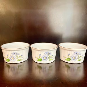 Puede incluir: Tres ramequines de cerámica blanca con un diseño floral. Cada ramequín presenta un exterior acanalado, un borde dorado y un patrón floral azul y verde. Los ramequines están dispuestos en fila sobre una superficie oscura.