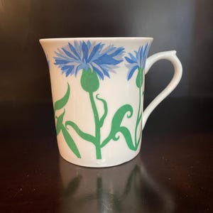 May include: White ceramic mug with a floral design. The mug features blue cornflower blossoms and green stems and leaves. The handle is white and curved. The mug is set against a dark background.