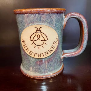 Puede incluir: Taza de cerámica con esmalte azul y marrón. Presenta un óvalo claro con un dibujo de luciérnaga y la palabra "FREETHINKER". El asa y el borde son marrones. Aproximadamente 10 cm de altura, ideal para bebidas calientes.
