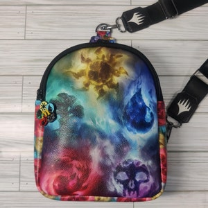 May include: A black leather crossbody bag with a colorful galaxy print. The bag features a zipper closure and a black adjustable strap with a metal buckle. The strap has a white logo that says "Magic: The Gathering".