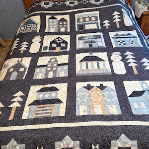 May include: A dark blue quilt with a repeating pattern of houses, trees, and snowmen in shades of blue, beige, and white. The quilt has a border with geometric shapes and is displayed on a wooden bed frame.
