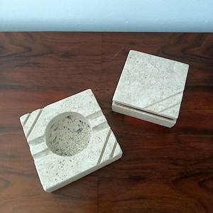 Könnte beinhalten: Ein quadratischer, cremefarbener Aschenbecher mit einer kreisförmigen Vertiefung und diagonalen Rillen. Neben dem Aschenbecher befindet sich eine passende quadratische Box mit Deckel. Beide Artikel bestehen aus einem gesprenkelten, porösen Material.