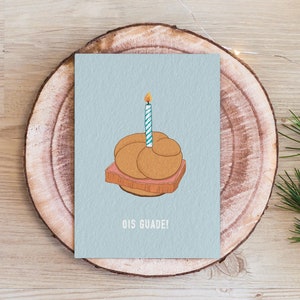 Könnte beinhalten: Eine hellblaue Grußkarte mit einer Cartoon-Illustration eines Brezels mit einer brennenden Kerze darauf. Der Text "Ois Guade!" ist unter dem Brezel gedruckt.