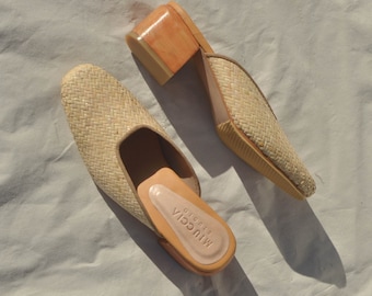 May include: A pair of woven straw heeled mules with a light brown wooden heel. The mules have a light brown woven upper and a light brown leather insole. The insole is labeled "Miucia Prada".
