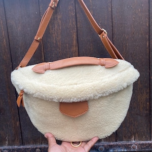 Puede incluir: Bolso de hombro color crema y esponjoso con detalles en cuero marrón. El bolso presenta una forma curva, un cierre de solapa y una correa de cuero marrón. Un pequeño parche de cuero marrón está en la parte delantera. El bolso tiene un anillo de metal dorado en la parte inferior.