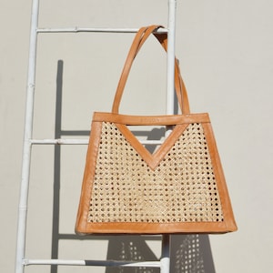 May include: A tan leather tote bag with a woven rattan panel and a V-shaped cutout. The bag has a leather strap and the word "MUCCIA" is printed on the front.