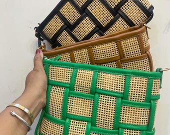 May include: Three woven rattan clutch bags in black, brown, and green. The bags have a rectangular shape with a woven pattern and leather trim. The green bag is held by a person's hand, showing a silver bracelet.