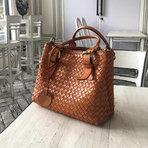 May include: A woven leather tote bag in a warm brown hue. The bag features two top handles, a shoulder strap, and a decorative tag. The bag has a rectangular shape with a woven pattern and is sitting on a white surface.