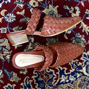 May include: A pair of brown woven leather heeled mules. The mules have a pointed toe and a low block heel. The brand name "MIUCCIA STUDIO" is printed on the insole of one of the mules.