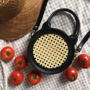 K&ouml;nnte beinhalten: Eine runde schwarze Ledertasche mit einem geflochtenen Stroheinsatz und einem oberen Henkel. Die Tasche hat einen langen Riemen und liegt auf einer karierten Tischdecke mit roten Tomaten.