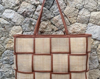 May include: A rectangular tote bag with a woven rattan design and brown leather trim. The bag features two brown leather handles and a structured shape. The woven pattern is a light beige colour, and the leather trim and handles are a medium brown.
