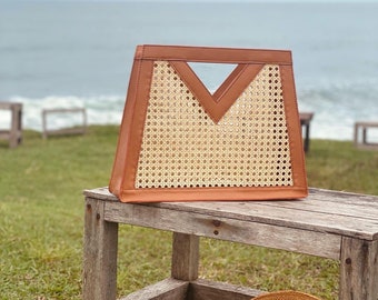 May include: A brown woven tote bag with leather trim sits on a wooden bench. The bag has a unique triangular cutout on the front. A straw hat sits on the bench below the bag.