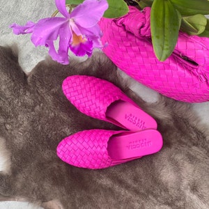 May include: Bright pink woven leather mules and a matching handbag are displayed on a textured, gray surface. A vibrant purple orchid and green leaves add a pop of color to the scene. The mules have the text "viccia studio" on the sole.