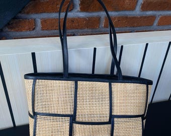 May include: A rectangular tote bag with a woven rattan body and black leather trim. The bag has two black handles and a structured design. The background includes a brick wall and a black surface.