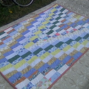 May include: A patchwork quilt featuring a mix of colours and patterns, including blue, yellow, green, and white. The quilt is made up of squares and rectangles, and it has a pink border.