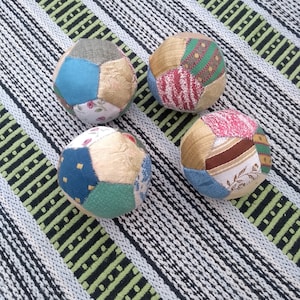 May include: Four patchwork fabric balls in various colours and patterns, including blue, green, red, and beige. The balls are arranged on a striped surface.