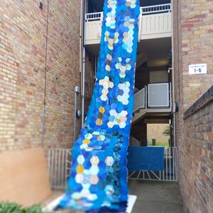 May include: A large blue and white patchwork quilt hangs from a balcony in a brick alleyway. The quilt is made of hexagonal fabric pieces and is hanging down to the ground.