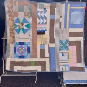 May include: A patchwork quilt with a variety of fabric patterns in shades of brown, blue, green, and pink. The quilt is displayed on a white metal rack, highlighting its handmade construction and intricate design. The quilt is a mix of geometric and floral patterns.