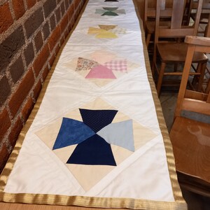 Könnte beinhalten: Ein weißer Tischläufer mit goldener Einfassung und einem Patchwork-Muster mit verschiedenen Farben und Mustern, darunter Rosa, Blau, Grün und Gelb. Der Läufer liegt auf einer Holzoberfläche mit Stühlen im Hintergrund.