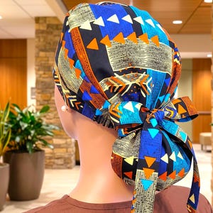 May include: A scrub cap featuring a vibrant geometric pattern in shades of blue, orange, black, and cream. The fabric displays repeating triangles and linear textures. The cap is secured at the back with a tied bow made from the same patterned fabric.