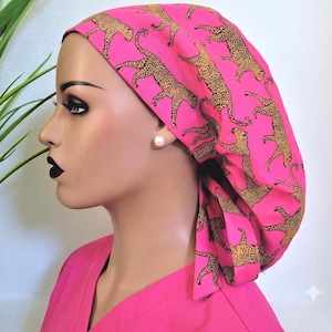 May include: A bright pink scrub cap featuring a repeating pattern of gold and black leopards. The cap is designed with a tie-back closure and is worn on a mannequin head, paired with a matching pink scrub top, showcasing a vibrant animal print.