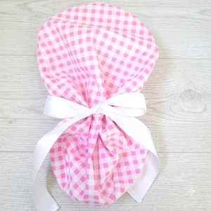 May include: A pink and white gingham patterned fabric hair accessory featuring a rounded, gathered shape. The fabric is cinched in the center with a smooth, solid white satin ribbon tied into a bow, with two long ribbon ends hanging downward.