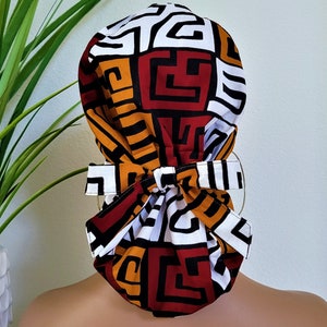 May include: A white, red, yellow, and black patterned surgical scrub hat with a tie at the back.