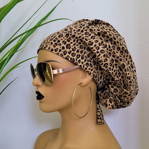 May include: A brown and tan leopard print surgical scrub hat with a tie. The hat is on a mannequin head wearing sunglasses and hoop earrings.