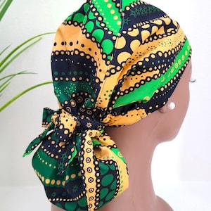 May include: A colorful patterned scrub cap in shades of green, yellow, and navy blue. The cap features a decorative bow at the back. The fabric has a repeating pattern of dots and circles. The cap is designed to be worn on the head.