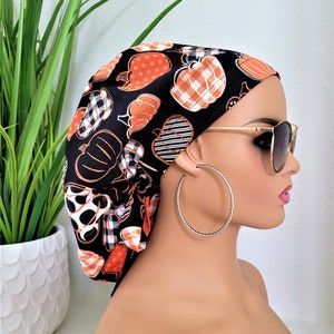 May include: Black and orange patterned fabric head wrap with a pumpkin design. The head wrap is worn by a person with sunglasses.